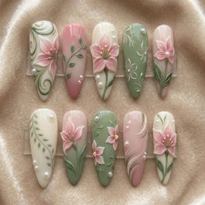 May include: A set of ten artificial nails with floral designs. Each nail features a different design, including pink and white flowers, green leaves, and pearl accents. The nails are painted in various colors, including pink, white, and green.