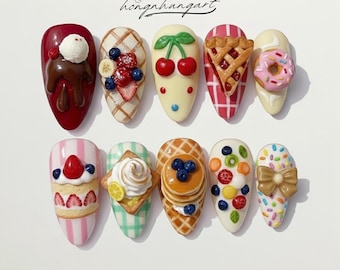 Könnte beinhalten: Zehn künstliche Fingernägel, die jeweils mit Miniatur-Lebensmitteldesigns verziert sind. Zu den Designs gehören ein Schokoladendessert, Obsttörtchen, Kirschen, ein Kuchen, ein Donut und Pfannkuchen. Der Text "hongnhungart" ist sichtbar. Die Nägel sind in verschiedenen Farben.