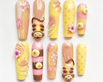 May include: A collection of ten long, artificial nails with various designs. The nails feature colors like yellow, pink, and brown, with decorative elements such as flowers, polka dots, and a monkey design. Some nails have a banana design.
