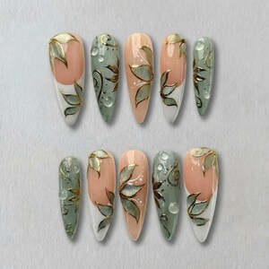 May include: A set of ten artificial nails with a floral design. The nails feature a combination of colors, including white, peach, and green, with gold leaf accents. Some nails have a water droplet effect.