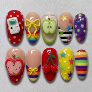 May include: A set of ten artificial nails with various designs. The designs include a red nail with a white game console, a nail with a yellow bow over a rainbow, and a nail with a green apple.