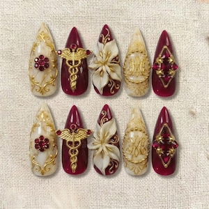 Op de afbeelding: Een set van tien lange, amandelvormige kunstnagels. De nagels hebben verschillende ontwerpen, waaronder gouden filigraan, rode edelstenen en witte bloemaccenten. Sommige nagels hebben een dieprode basis, andere zijn crèmekleurig.
