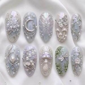 May include: A set of ten almond-shaped press-on nails featuring an iridescent finish. Designs include silver metallic accents, white floral reliefs, a crescent moon, a butterfly, mushrooms, a bee, a dragonfly, and small embedded rhinestones.