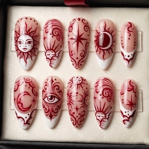 May include: A set of ten almond-shaped false nails. Each nail features a design in burgundy on a nude base, with white tips. Designs include suns, moons, stars, and celestial patterns. The nails are arranged in a black box.