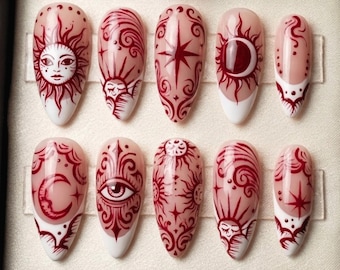 Puede incluir: Un conjunto de diez uñas postizas en forma de almendra. Cada uña presenta un diseño burdeos sobre una base nude, con puntas blancas. Los diseños incluyen soles, lunas, estrellas y patrones celestiales. Las uñas están dispuestas en una caja negra.