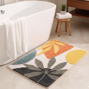 May include: A rectangular bath mat with a botanical design. The mat features a white background with colorful leaf and geometric shapes in shades of grey, orange, yellow, and teal. The mat is placed in front of a white bathtub.