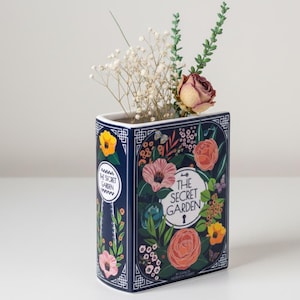 Könnte beinhalten: Eine marineblaue Keramikvase in Buchform mit floralen Illustrationen und dem Text "The Secret Garden". Die Vase enthält getrocknete Blumen und Grünzeug. Das Design umfasst rosa, orange und gelbe Blumen.