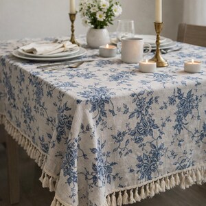 Peut inclure: Une nappe en lin avec un motif floral bleu et une bordure à franges. La table est dressée avec des assiettes blanches, des bougies dans des bougeoirs en laiton, un vase de fleurs blanches et une tasse blanche. La nappe est drapée sur une table en bois.