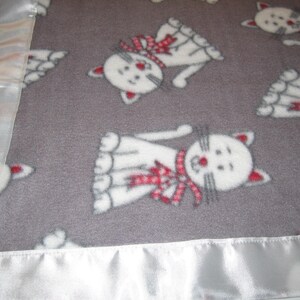 May include: A gray fleece blanket with a white satin border. The blanket features a repeating pattern of white cats with red bows.