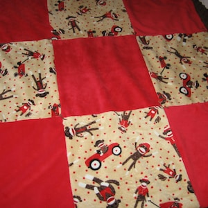 May include: A red and beige patchwork quilt with a pattern of monkeys wearing red socks and driving red toy cars. The quilt is made of soft, plush fabric.