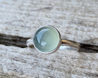 Puede incluir: Un anillo de plata con una piedra preciosa cabujón redonda, lisa y de color verde pálido engastada en un bisel sencillo. La banda es de plata pulida y la piedra tiene una superficie brillante y reflectante que capta la luz.