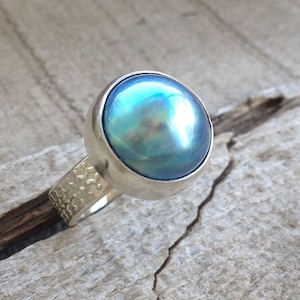 May include: A silver ring with a blue iridescent pearl set in a bezel setting. The band of the ring has a textured design.