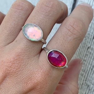 May include: Two silver rings featuring oval-shaped faceted stones. One ring displays a pale, iridescent, light-reflecting stone, while the other ring features a vibrant, deep magenta-coloured stone with a faceted surface texture.