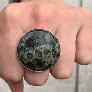 Könnte beinhalten: Ein silberner Ring mit einem großen, runden, grün-schwarzen Stein. Der Stein hat ein marmoriertes Muster.