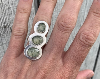May include: A silver ring with three green stones set in a unique, asymmetrical design.