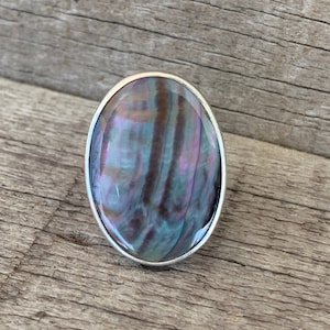 May include: A silver ring with an oval-shaped abalone shell inlay. The shell has a iridescent blue, green, and purple color.