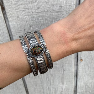 Könnte beinhalten: Drei silberne Armbänder mit aufwendigen Designs, übereinander auf einem Handgelenk getragen. Das mittlere Armband hat einen großen, ovalen Opal-Stein mit grünen, roten und orangefarbenen Farbtönen.