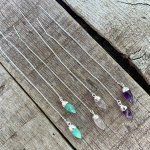 May include: Six pairs of silver chain earrings with different colored gemstone tips. The gemstones are green, pink, and purple.