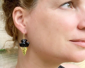 May include: A close-up of a gold hoop earring with a black, textured top and a gold arrowhead-shaped bottom. The earring is worn on a person's ear. The background is a wooden fence.