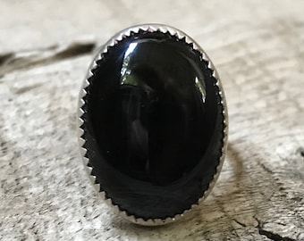 May include: A ring featuring a large, smooth, oval-shaped black stone set in a silver-toned metal bezel. The metal setting has a decorative, notched, or serrated edge surrounding the perimeter of the polished black gemstone.