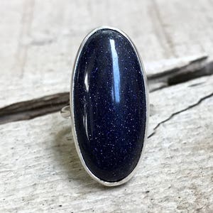 Könnte beinhalten: Ein silberner Ring mit einem dunkelblauen, ovalen Edelstein. Der Edelstein hat eine funkelnde, gesprenkelte Textur.