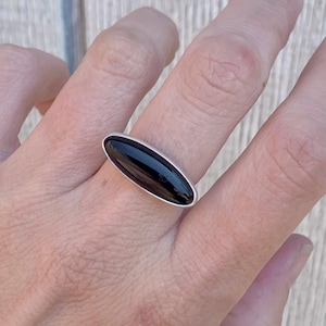 May include: A silver ring featuring a large, oval, black stone. The stone is set in a polished silver band. The ring is shown on a finger, highlighting the sleek design and the contrast between the dark stone and the silver.