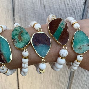 May include: Five bracelets with white beads and a large green and brown stone pendant on each. The bracelets are stacked on a person's wrist.