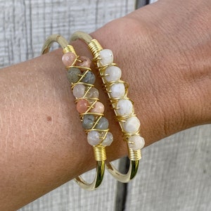 May include: Two gold wire wrapped bracelets with different colored stones. One bracelet has white stones and the other has pink, white, and gray stones.