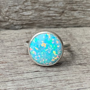 Puede incluir: Un anillo de plata con una piedra redonda de ópalo azul. El ópalo tiene una base blanca con destellos iridiscentes verdes, amarillos y rosados.