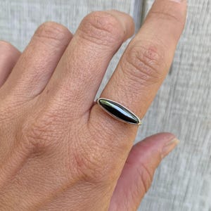 Puede incluir: Un anillo de plata con una piedra negra pulida y alargada. El anillo tiene una banda fina y sencilla, con la piedra oscura engastada en un estilo minimalista. El anillo se muestra en un dedo sobre un fondo de madera clara.