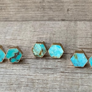 May include: A collection of six turquoise hexagon stud earrings with gold-colored edges. The earrings are arranged in three pairs, each showcasing the unique patterns and colors of the turquoise stones.