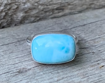May include: A silver ring with a rectangular, pale blue gemstone. The gemstone has a smooth, polished surface and is set in a simple silver band. The ring is displayed on a weathered wooden surface.