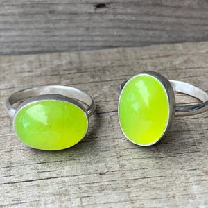 Könnte beinhalten: Zwei Silberringe mit ovalen, grünen Edelsteinen. Die Ringe liegen auf einer Holzoberfläche.