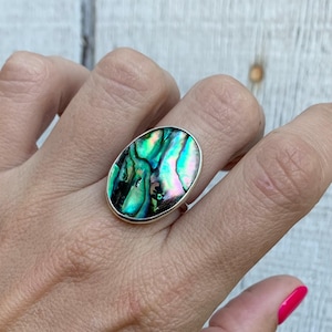 May include: Oval-shaped ring featuring a vibrant abalone shell with iridescent green, blue, and pink hues. The shell is set in a silver-toned metal band. A unique piece of jewelry.