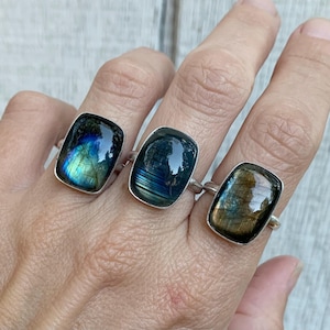 May include: Three silver rings featuring rectangular labradorite gemstones. The left ring displays a vibrant blue and green flash, the center ring shows deep blue horizontal striations, and the right ring exhibits a warm golden brown iridescence.