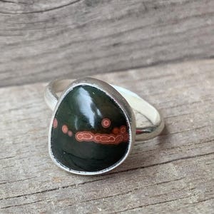 Könnte beinhalten: Ein silberner Ring mit einem dunkelgrünen Edelstein mit roten und orangefarbenen Flecken. Der tropfenförmige Stein ist in eine silberne Fassung gefasst. Der Ring wird auf einer Holzoberfläche gezeigt.