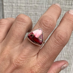 May include: A silver ring with a triangular, multi-colored stone. The stone features a mix of deep red, pink, and gold hues. The ring's setting is a simple, polished silver band, showcasing the vibrant stone.