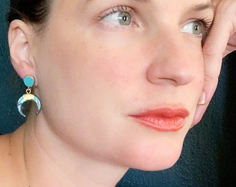 May include: A pair of gold crescent moon earrings with turquoise enamel accents. The earrings are dangling and have a small turquoise circle at the top.