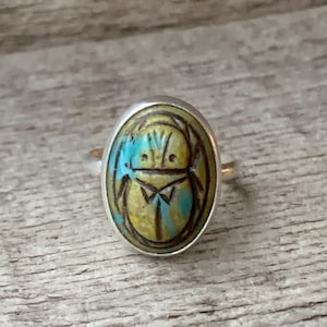 May include: Silver ring featuring an oval-shaped, painted scarab beetle design. The beetle is depicted in shades of yellow, blue, and black. The ring has a simple, elegant band.