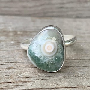 Könnte beinhalten: Ein Silberring mit einem einzigartigen, unregelmäßig geformten Stein. Der Stein hat ein zentrales weißes und hellbraunes Wirbelmuster, umgeben von einem seegrünen und weißen gesprenkelten Design. Der Ring hat ein schlichtes, poliertes Silberband.