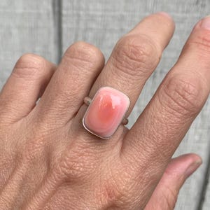 Peut inclure: Bague rectangulaire en argent avec une pierre rose. La pierre présente un dégradé de rose, plus clair sur les bords et plus foncé au centre. La bague est portée au doigt.