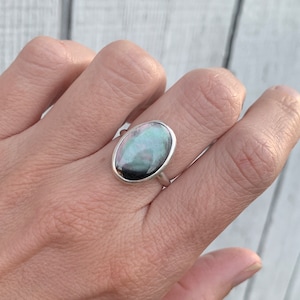 May include: A silver ring featuring an oval-shaped, iridescent gemstone. The stone displays a mix of green, blue, and black hues. The ring's band is a simple, polished silver.