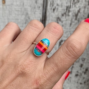 May include: A silver ring featuring an oval-shaped inlay composed of horizontal stripes in vibrant shades of turquoise, orange, hot pink, and dark purple. The metal band is smooth and polished, holding the multi-colored stone setting securely in place.