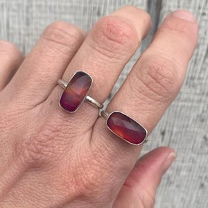 May include: Two silver rings, each featuring a rectangular, faceted gemstone. The gemstones display a gradient of colors, ranging from deep red to orange and purple. The rings are set on a hand, showcasing the jewelry's design and color variations.