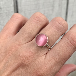 May include: A silver ring with a pink round gemstone. The ring is on a person's finger.