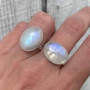 Puede incluir: Dos anillos de plata con piedras de luna ovaladas. Las piedras son iridiscentes con tonos de azul y blanco.
