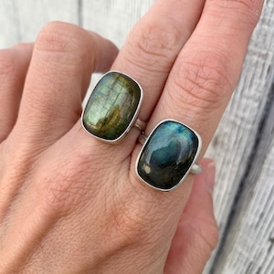 May include: Two silver rings with green and blue labradorite gemstones. The rings are on a person's finger.