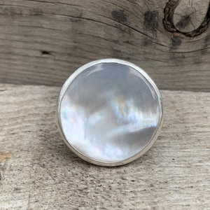 May include: A silver ring featuring a large, round, white mother-of-pearl stone.