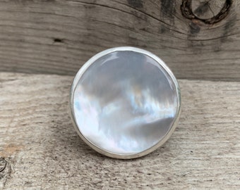 May include: A silver ring with a large, round, white mother-of-pearl stone.