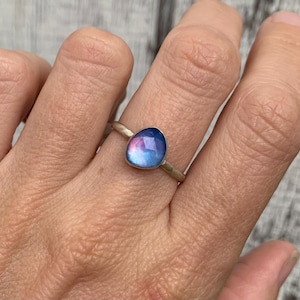 May include: A silver ring featuring a single, faceted, teardrop-shaped stone with a smooth, polished surface. The stone displays a gradient of iridescent blue and pink hues that shift in color across its surface, set in a simple metal band.